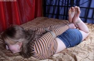 White girl is left on a bed while hogtied in a long sleeved shirt and cutoffs on galphoto.com